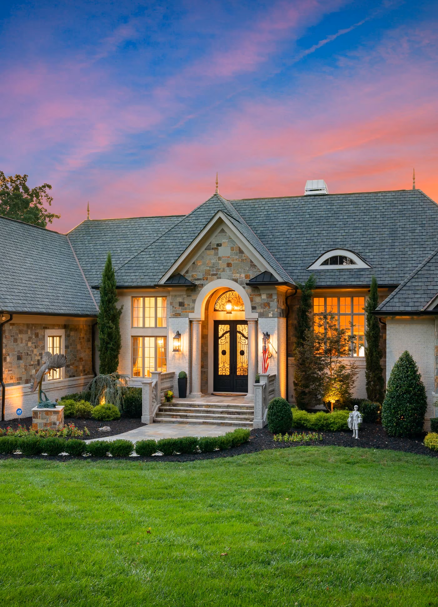 A stone house lit at dusk, its arched entry centred on the lawn.